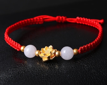 May include: A red string bracelet with two white beads and a gold lotus flower charm.