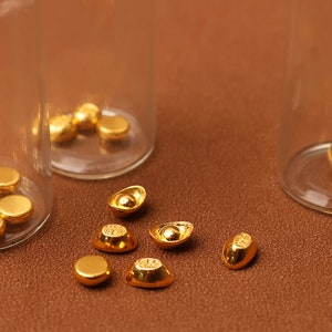 May include: Close-up of gold-colored ingots and round coins, some inside clear glass vials. The objects are scattered on a brown surface. The ingots have a boat-like shape, and the coins are flat and circular.