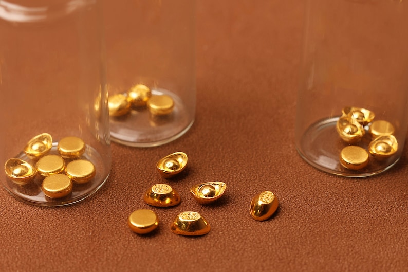 May include: Close-up of gold-colored ingots and round coins, some inside clear glass vials. The objects are scattered on a brown surface. The ingots have a boat-like shape, and the coins are flat and circular.