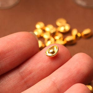 May include: Close-up of several small, gold-colored, ingot-shaped beads. One bead is held between two fingers, with others scattered in the background. The beads have a shiny, metallic finish and a small, rounded shape.