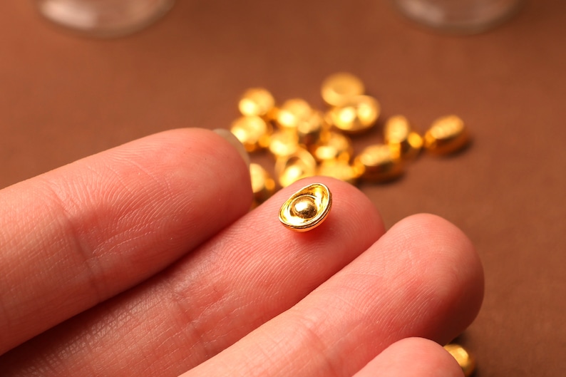May include: Close-up of several small, gold-colored, ingot-shaped beads. One bead is held between two fingers, with others scattered in the background. The beads have a shiny, metallic finish and a small, rounded shape.