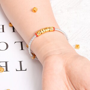 May include: A white leather bracelet with gold letter beads spelling "LIKE" on a person's wrist.
