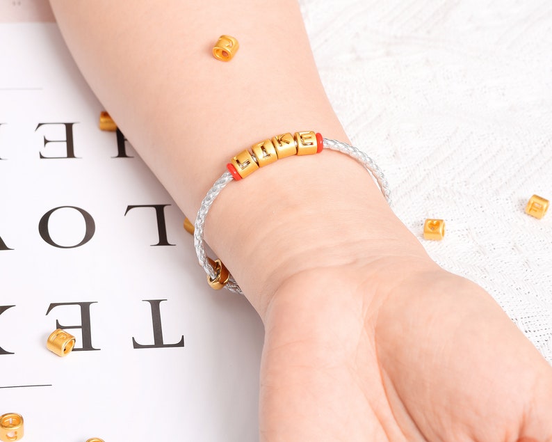 May include: A white leather bracelet with gold letter beads spelling "LIKE" on a person's wrist.