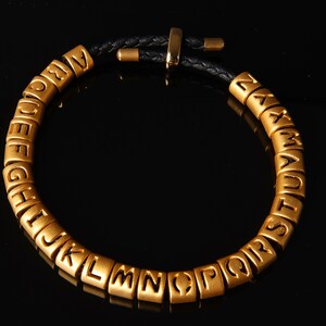 May include: A gold alphabet bracelet with black leather cord. Each letter is a separate bead.