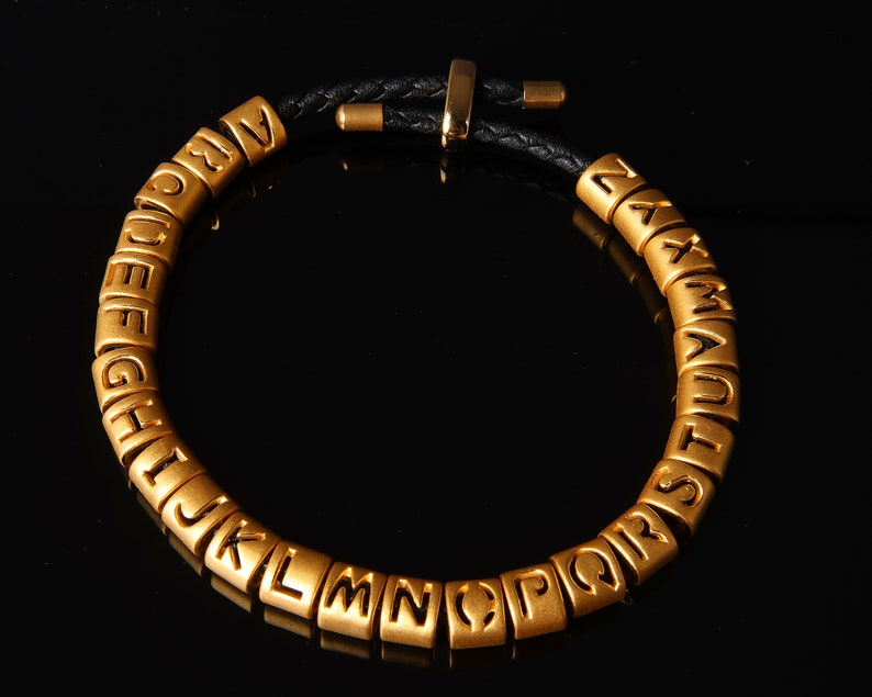May include: A gold alphabet bracelet with black leather cord. Each letter is a separate bead.