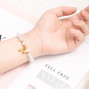 May include: White beaded bracelet with a gold charm featuring three small bells. The bracelet is on a person's wrist.