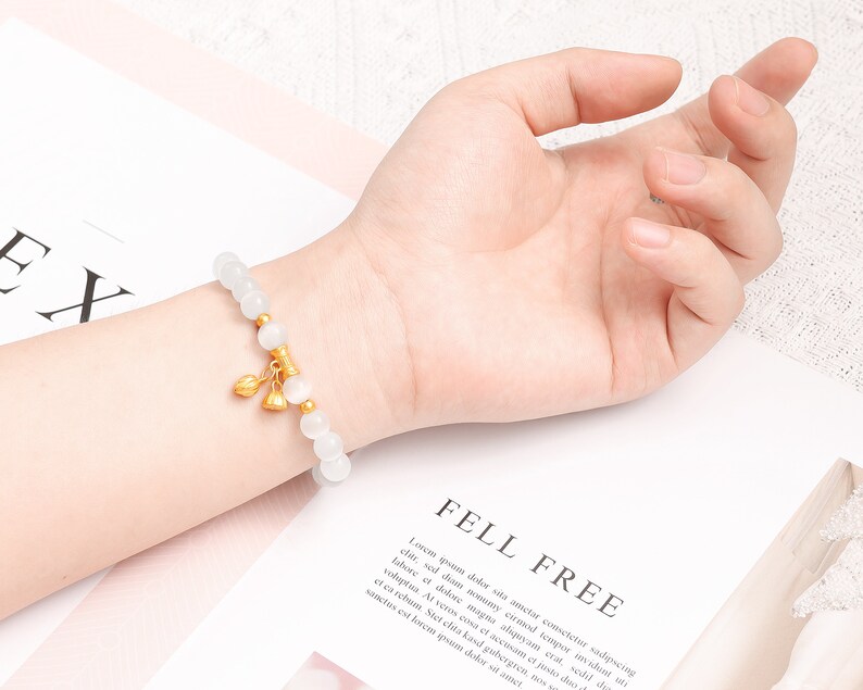 May include: White beaded bracelet with a gold charm featuring three small bells. The bracelet is on a person's wrist.