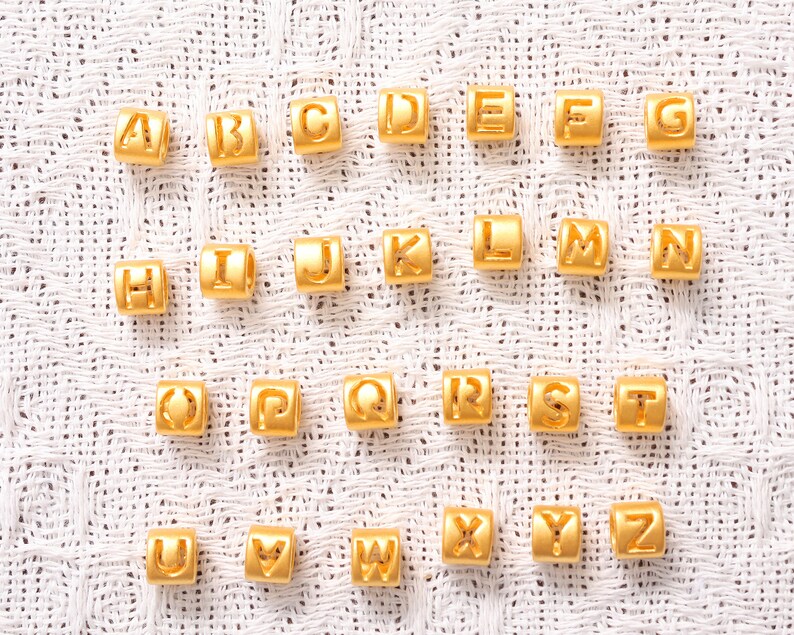 May include: Gold alphabet letter beads, each letter is a separate bead. The beads are arranged in alphabetical order on a white fabric background.
