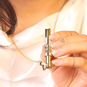 May include: A gold and silver necklace with a pendant that is a small, cylindrical tube with a decorative design. The tube is open at one end and has a small, clear stone at the other end.