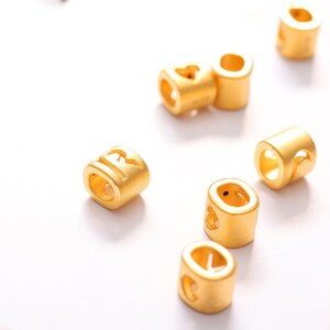 May include: A collection of gold-colored metal beads with different letters and symbols engraved on them. The beads are designed to be used for jewelry making or other craft projects.