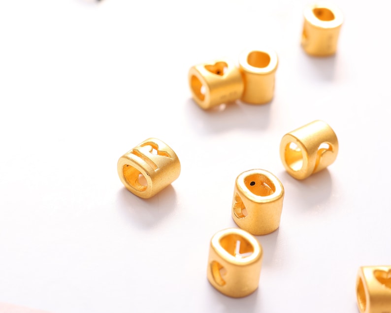May include: A collection of gold-colored metal beads with different letters and symbols engraved on them. The beads are designed to be used for jewelry making or other craft projects.