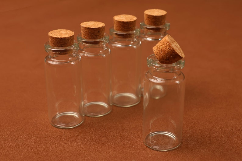 May include: Clear glass bottles with cork stoppers. The bottles are cylindrical with a narrow neck and a wider base. The corks are a light brown color. One cork is removed from its bottle. The background is a solid brown color.