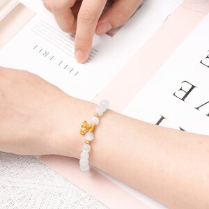 May include: A white beaded bracelet with a gold charm. The charm is a small, stylized design of a bird.