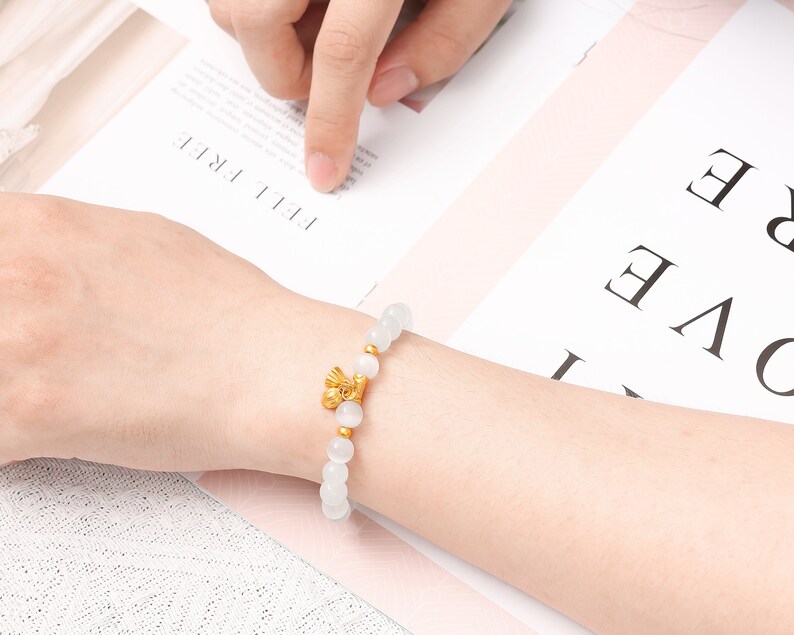 May include: A white beaded bracelet with a gold charm. The charm is a small, stylized design of a bird.
