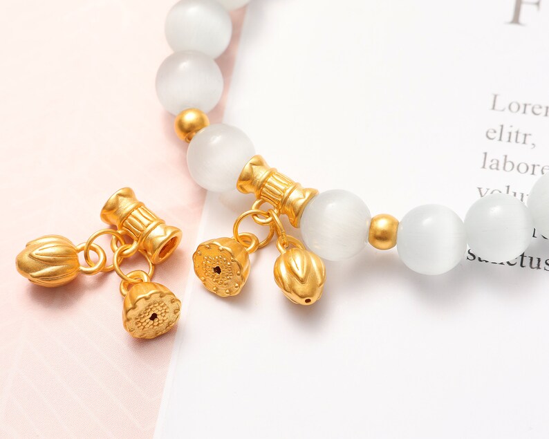 May include: A white gemstone bracelet with gold accents and charms. The charms are shaped like lotus flowers and have a detailed design.