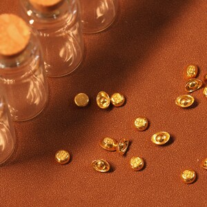 May include: A collection of small, gold-colored ingots and round coins scattered on a brown surface. Three clear glass bottles with cork stoppers are arranged in the background. The lighting creates shadows, highlighting the textures and shapes.