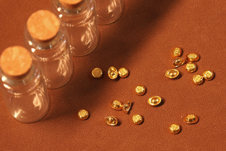 May include: A collection of small, gold-colored ingots and round coins scattered on a brown surface. Three clear glass bottles with cork stoppers are arranged in the background. The lighting creates shadows, highlighting the textures and shapes.