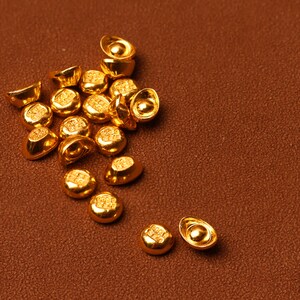 May include: A collection of small, gold-colored ingots, each with a unique shape and embossed design. The ingots are scattered on a textured, brown surface, creating a warm and inviting visual.