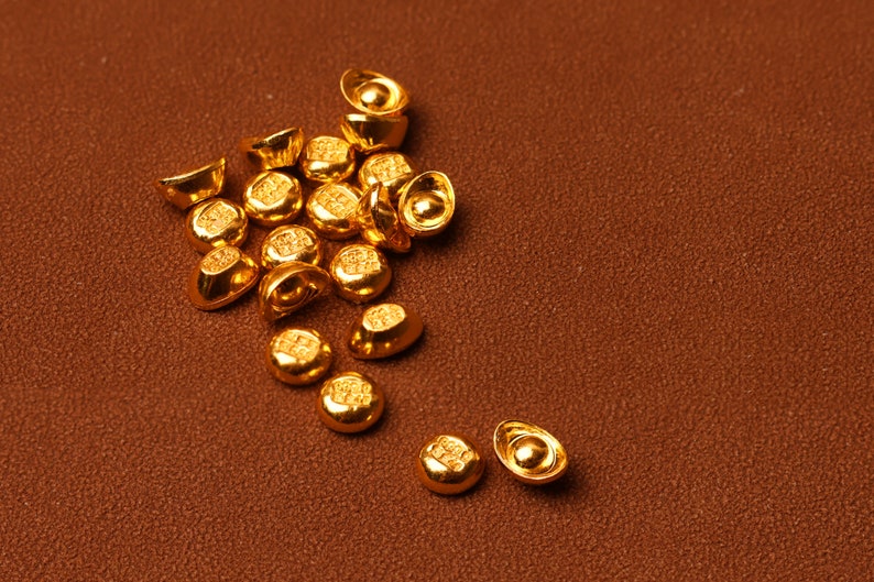 May include: A collection of small, gold-colored ingots, each with a unique shape and embossed design. The ingots are scattered on a textured, brown surface, creating a warm and inviting visual.