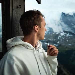 May include: A person wearing a white hoodie with a black chain and a silver vape pen. The person is looking out of a window at a snowy mountain range.