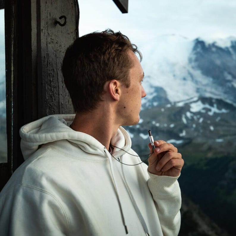 May include: A person wearing a white hoodie with a black chain and a silver vape pen. The person is looking out of a window at a snowy mountain range.