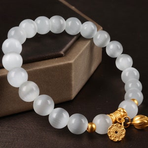May include: White gemstone bracelet with gold accents and a flower charm. The bracelet is made of round beads and has a delicate, feminine look.