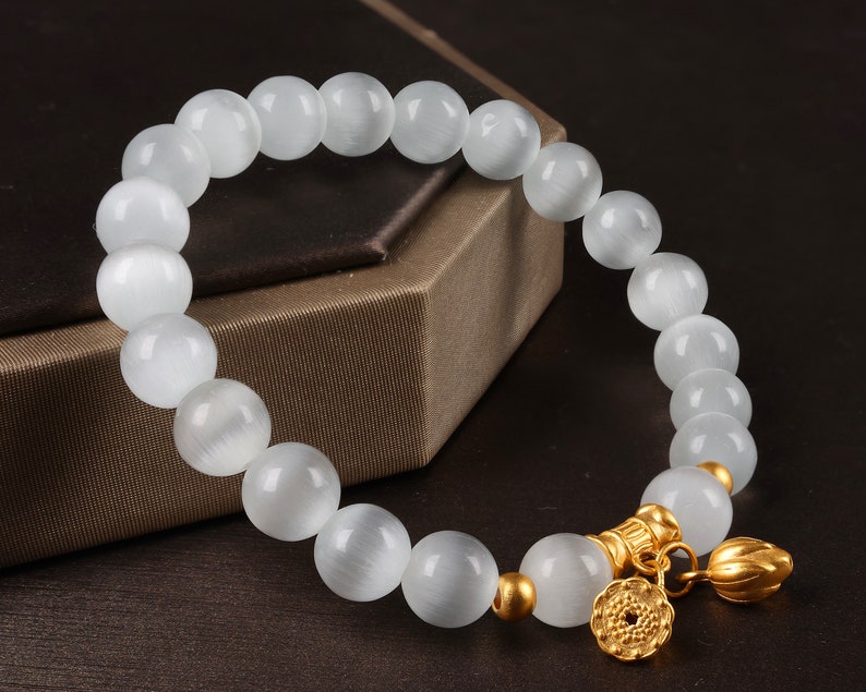 May include: White gemstone bracelet with gold accents and a flower charm. The bracelet is made of round beads and has a delicate, feminine look.