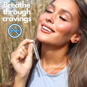 May include: A woman wearing a light blue shirt holds a silver necklace with a small silver cylinder pendant. The pendant has a design of a cigarette with a line through it. The text "Breathe through cravings" is in the top left corner of the image.