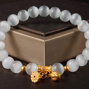 May include: A white gemstone bracelet with gold accents. The bracelet features a chain with two gold charms, one with a floral design and the other with a lotus flower design.