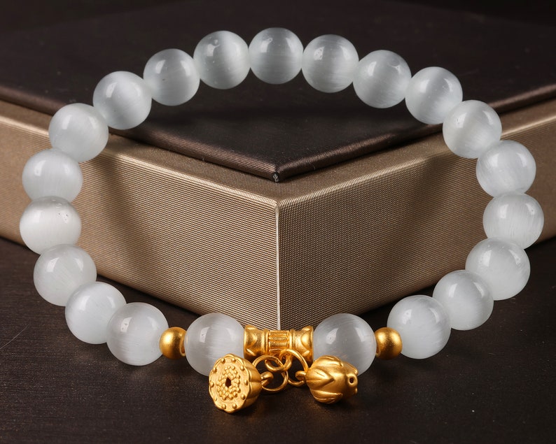 May include: A white gemstone bracelet with gold accents. The bracelet features a chain with two gold charms, one with a floral design and the other with a lotus flower design.