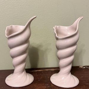 May include: Two matching, off-white ceramic vases. Each vase has a spiral-shaped stem and a flared top with a curved, pointed edge. The vases are displayed on a dark wooden surface.