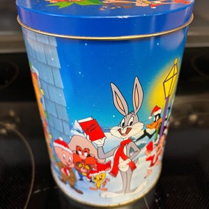 Puede incluir: Una lata de metal azul con un tema navideño con Bugs Bunny, Pato Lucas, Piolín y otros personajes de Looney Tunes. La lata tiene una bufanda roja y blanca a rayas alrededor del cuello de Bugs Bunny y un libro rojo que dice "Christmas Carols".