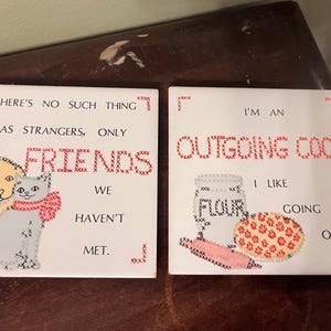 May include: Two square white ceramic tiles featuring cross-stitch style designs. The left tile shows a dog and cat with the text, THERE'S NO SUCH THING AS STRANGERS, ONLY FRIENDS WE HAVEN'T MET. The right tile shows a flour jar and pie with the text, I'M AN OUTGOING COOK I LIKE GOING OUT.