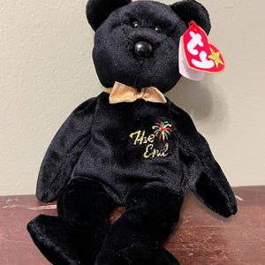 Puede incluir: Oso de peluche negro con pajarita dorada y el texto bordado "The End" en hilo dorado. El oso tiene una etiqueta roja con texto blanco y una estrella. El oso está sentado sobre una superficie de madera.