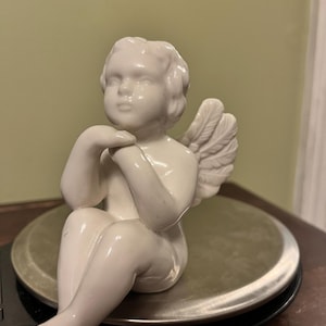 May include: A white ceramic cherub figurine with detailed wings, seated with hands near the face. The cherub is on a silver scale, set against a neutral background. The figurine is a decorative item.
