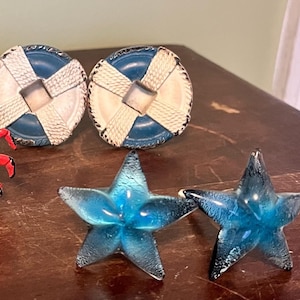 Peut inclure: Une collection d'objets décoratifs sur le thème nautique. Deux boutons de tiroir en forme de bouée de sauvetage bleues et blanches, et deux étoiles de mer en verre bleu. Les objets sont disposés sur une surface en bois foncé.