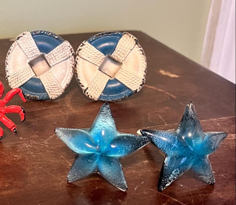 以下が含まれることがあります： 航海をテーマにした装飾品のコレクション。青と白の救命浮き輪型の引き出しつまみ2つと、青いガラスのヒトデ2つ。アイテムは濃い木製の表面に配置されています。