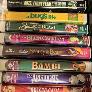 Könnte beinhalten: Ein Stapel VHS-Kassetten mit klassischen Disney-Animationsfilmen, darunter Cinderella, Buzz Lightyear of Star Command, Ein Leben als Käfer, Die Schöne und das Biest, Der schwarze Kessel, Die Schöne und das Biest, Bambi, Die Aristocats und Die Aristocats.