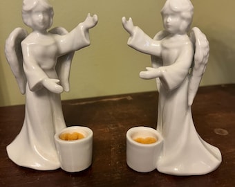 Puede incluir: Dos figuras de ángeles de cerámica blanca con los brazos y las alas extendidos. Cada ángel sostiene un pequeño portavelas redondo con una vela amarilla en su interior. Los ángeles están sobre una superficie marrón oscuro.