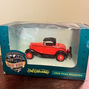 May include: A red 1932 Ford Roadster model car with black accents and a black convertible top, displayed in a clear-windowed box. The box has the text "Authentic Railway Designs" and "Ertl Collectibles".