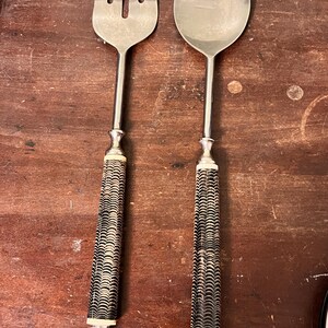 May include: A silver serving fork and spoon set. The handles are cylindrical with a black and white wave pattern. The fork has three tines, and the spoon has a rounded bowl. The set is likely for serving salads or other dishes.