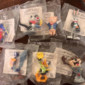 Op de afbeelding: Verzameling Looney Tunes verzamelfiguren, elk verzegeld in een plastic zak. De figuren omvatten personages als Bugs Bunny, Sylvester en de Tasmanian Devil. Elke figuur staat op een kleine kartonnen achtergrond.