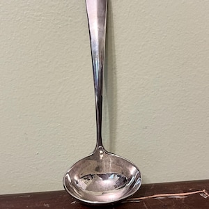 Könnte beinhalten: Eine silberne Schöpfkelle mit langem Stiel und rundem Napf. Die Schöpfkelle besteht aus glänzendem, reflektierendem Metall, und der Griff hat eine glatte, polierte Oberfläche. Die Schöpfkelle ist zum Servieren von Suppen, Saucen oder anderen Flüssigkeiten bestimmt.