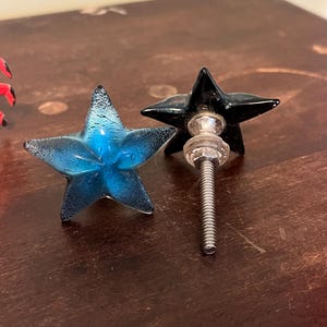 以下が含まれることがあります： 星型のキャビネットノブが2つ。1つは半透明の青色で、表面にテクスチャがあり、もう1つは黒色です。各ノブには、取り付け用の金属ネジが付いています。家具用の装飾金具。