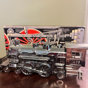 May include: A shiny chrome toy train with intricate wheel detailing and a small caboose. The train is positioned in front of a box with a train illustration. The train sits on a wooden surface.