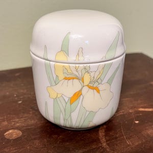 May include: A white ceramic jar with a lid, featuring a floral design of yellow and orange irises with green leaves. The jar has a rounded shape and sits on a wooden surface.