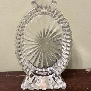 May include: Clear glass oval-shaped picture frame with a scalloped edge and a fan-like design in the center. The frame stands on a decorative base. The frame is made of clear glass.