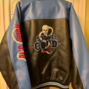 以下が含まれることがあります： 背面にスカルとヘビの刺繍グラフィックと「STONE COLD」「Austin 3:16」の文字が入った、青と黒のフェイクレザー製バーシティジャケット。袖は青色で、赤い文字とヘビの刺繍が施されており、襟と袖口はストライプ柄になっています。