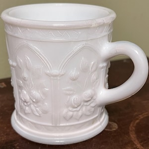 May include: White milk glass mug with a handle. The mug features a raised floral design with roses and leaves. The rim and base have a decorative border. The mug is a vintage style and is suitable for hot or cold drinks.