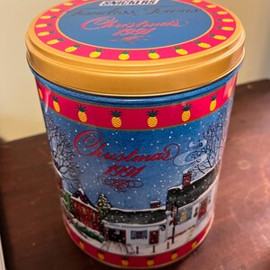 Könnte beinhalten: Eine Vintage-Snickers-Dose mit einem blauen und roten Design, das eine verschneite Winterszene mit einem Haus und dem Text "Christmas 1991" zeigt. Der Deckel ist golden mit einem roten und gelben Design mit Ananas und dem Text "Snickers Timeless Treats Christmas 1991".